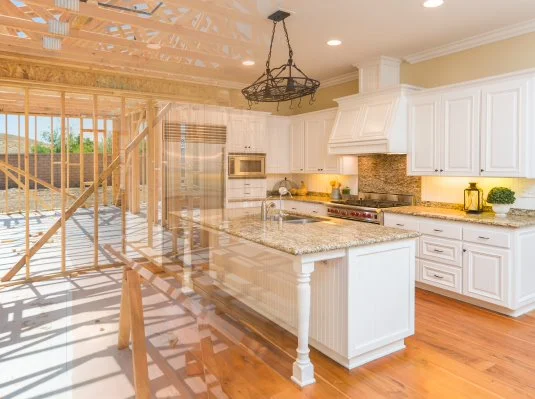 A modern kitchen with white cabinets and granite countertops is overlaid with an image of its wooden frame under construction, highlighting transformation.