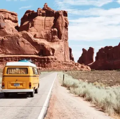 A yellow van travels down a deserted road in a vast desert landscape surrounded by towering red rock formations under a blue sky, evoking a sense of adventure.