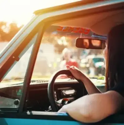 A person sits in a blue car, hand on the steering wheel, gazing at a hazy, sunlit scene outside. The ambiance is relaxed and warm.