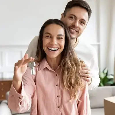 Smiling couple in a bright room, embracing, as the woman holds a set of keys. A moving box is visible nearby, suggesting a new home.