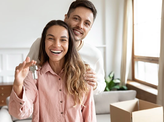 Smiling couple in a bright room, embracing, as the woman holds a set of keys. A moving box is visible nearby, suggesting a new home.