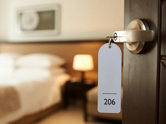 A hotel room door is slightly open, revealing a cozy interior with a neatly made bed and a warm-lit bedside lamp. A tag hangs from the door labeled "206."
