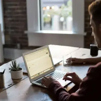 A man sits at a table, focused on his laptop, with a neutral background.