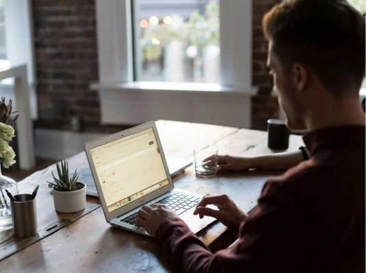 A man sits at a table, focused on his laptop, with a neutral background.