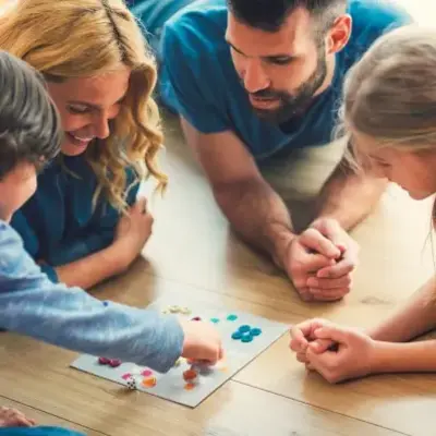 A family of four happily plays a board game on a wooden floor. Parents and children are engaged, smiling, fostering a warm, joyful atmosphere.