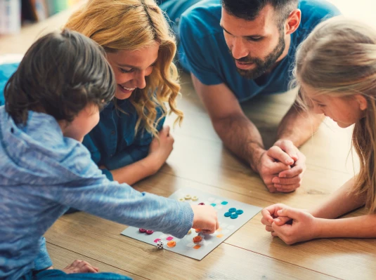A family of four happily plays a board game on a wooden floor. Parents and children are engaged, smiling, fostering a warm, joyful atmosphere.