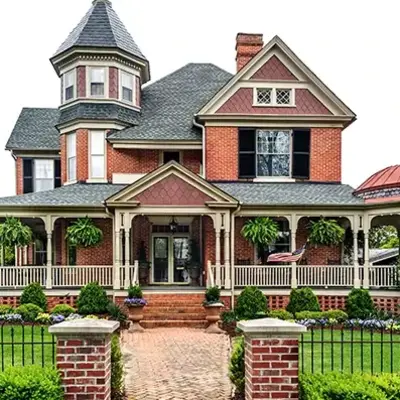 Victorian-style brick house with a wraparound porch, ornate turret, and lush landscaping. Features hanging plants, a manicured lawn, and a vintage charm.