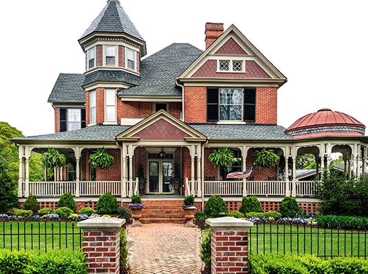 Victorian-style brick house with a wraparound porch, ornate turret, and lush landscaping. Features hanging plants, a manicured lawn, and a vintage charm.