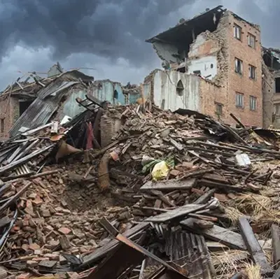 Collapsed brick buildings under a dark, stormy sky. Piles of debris and rubble are scattered across the foreground, conveying destruction and chaos.