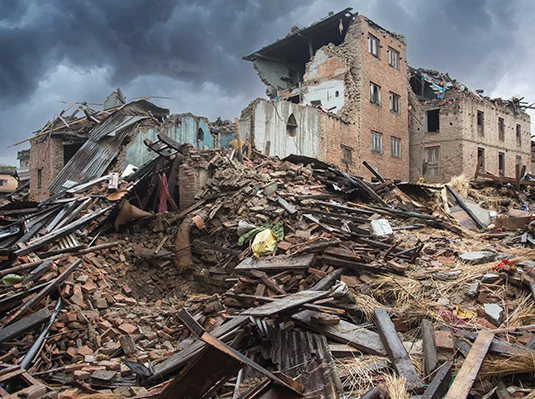 Collapsed brick buildings under a dark, stormy sky. Piles of debris and rubble are scattered across the foreground, conveying destruction and chaos.