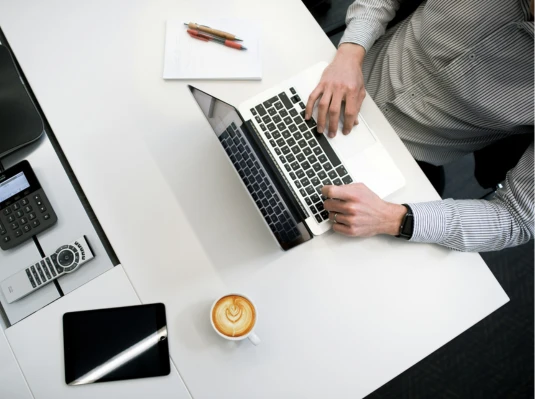 A man seated at a desk, working on a laptop with a cup of coffee beside him.