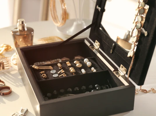 An open black jewelry box on a table holds gold and pearl earrings, a necklace, and bracelets. Perfume and more accessories surround it, creating a luxurious, elegant setting.