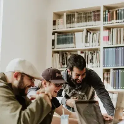 Three people are laughing and working together on laptops in a library filled with bookshelves. The atmosphere is collaborative and joyful.