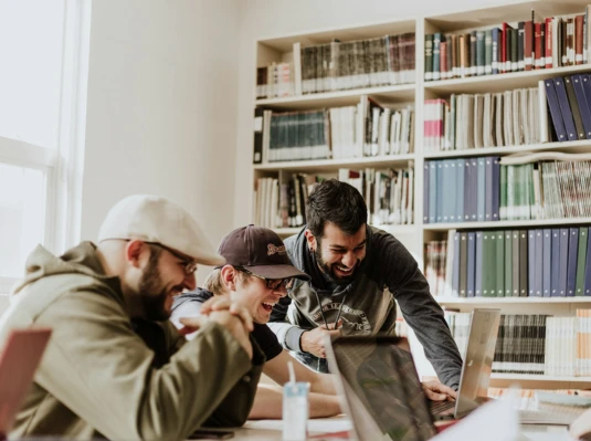 Three people are laughing and working together on laptops in a library filled with bookshelves. The atmosphere is collaborative and joyful.
