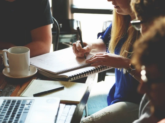 A group seated around a table with coffee, a laptop, and notebooks, engaged in discussion. A person writes notes, conveying focus and collaboration.