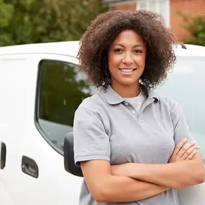 A woman in a gray shirt stands beside a white van, smiling and looking towards the camera.