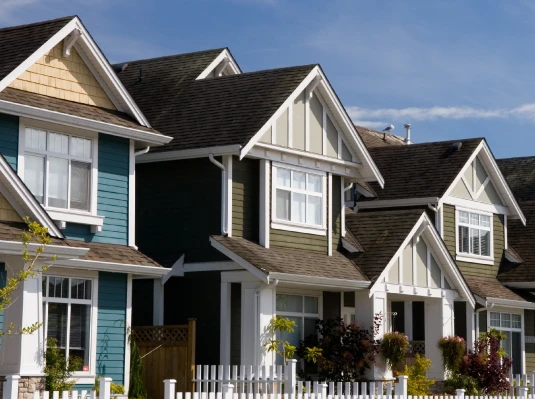 Suburban neighborhood with colorful, well-maintained houses featuring pitched roofs and white picket fences under a clear blue sky, conveying a charming, peaceful atmosphere.