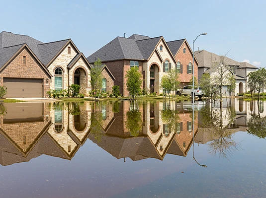 Row of suburban homes reflected in floodwaters, highlighting the impact of flooding. The scene is calm with clear skies, emphasizing the severity.