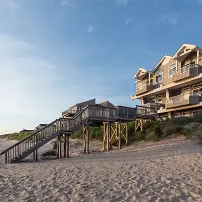 A large beach house with wooden stairs sits on sandy dunes under a clear blue sky. The scene conveys a serene and inviting coastal atmosphere.