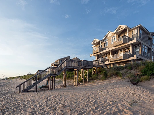 A large beach house with wooden stairs sits on sandy dunes under a clear blue sky. The scene conveys a serene and inviting coastal atmosphere.