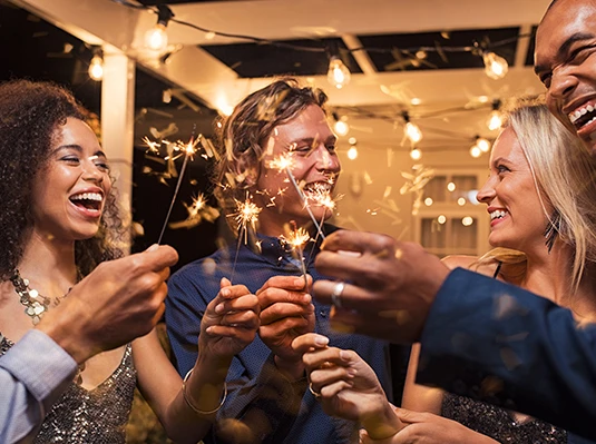 A group of people smiling and holding sparklers at a night party under string lights. The atmosphere is festive and joyful, highlighting celebration.