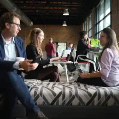 Casual office setting with three people chatting on a patterned bench, laptops open. Others work at high tables near large windows, creating a collaborative vibe.