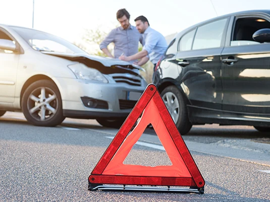 A red warning triangle is placed on the road in front of two cars that appear to have collided. Two men are inspecting the damage, looking concerned.