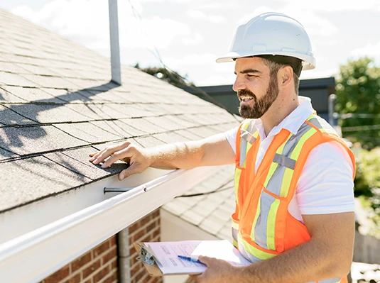 A construction worker in a white hard hat and orange safety vest inspects a roof, smiling while holding a clipboard. It's a sunny day, conveying positivity.