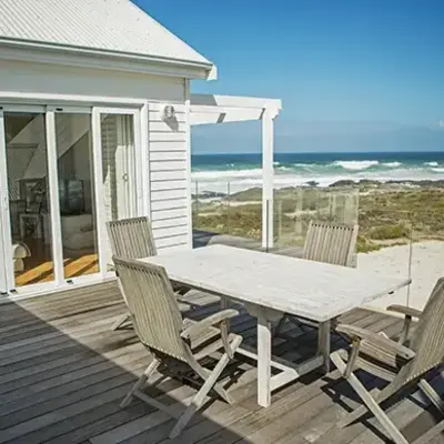 A wooden deck with a white table and four chairs overlooks a beach. Glass doors lead to a cozy interior. The scene conveys calmness and relaxation.