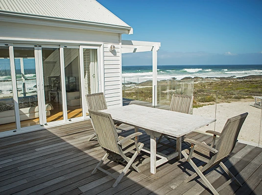 A wooden deck with a white table and four chairs overlooks a beach. Glass doors lead to a cozy interior. The scene conveys calmness and relaxation.
