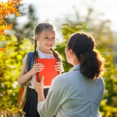 A young girl holding a red notebook smiles at a woman in a garden setting. The bright, sunny background enhances the joyful, warm atmosphere.