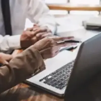 Two people in business attire are seated at a desk, viewing a laptop. One gestures with a pen, indicating collaboration and focus. Papers and a plant are visible.