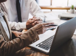 Two people in business attire are seated at a desk, viewing a laptop. One gestures with a pen, indicating collaboration and focus. Papers and a plant are visible.