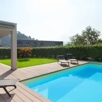 Modern patio with a wooden deck, bright blue pool, and lounge chairs. Green hedge and small tree provide a serene, inviting atmosphere.