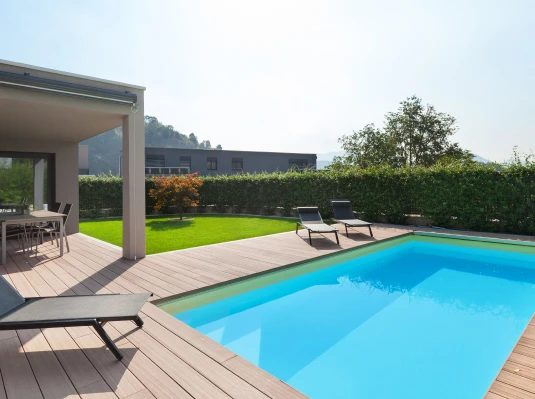 Modern patio with a wooden deck, bright blue pool, and lounge chairs. Green hedge and small tree provide a serene, inviting atmosphere.