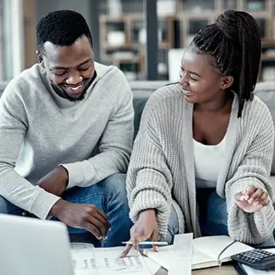 A smiling couple sits on a sofa reviewing documents, pointing at papers with a laptop nearby. The scene conveys teamwork and a relaxed, supportive atmosphere.