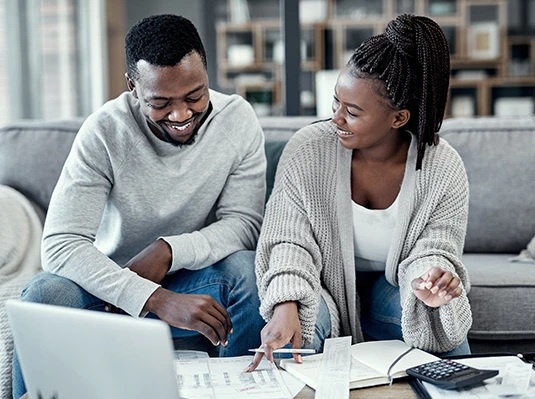 A smiling couple sits on a sofa reviewing documents, pointing at papers with a laptop nearby. The scene conveys teamwork and a relaxed, supportive atmosphere.