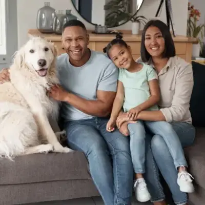 A joyful family of four, including a large fluffy dog, sits on a cozy grey sofa. The parents and child, all smiling, exude warmth and happiness.
