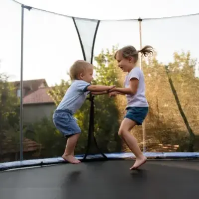 Two young children joyfully bounce on a trampoline, holding hands. They are surrounded by trees and houses on a sunny day, conveying a playful, carefree mood.