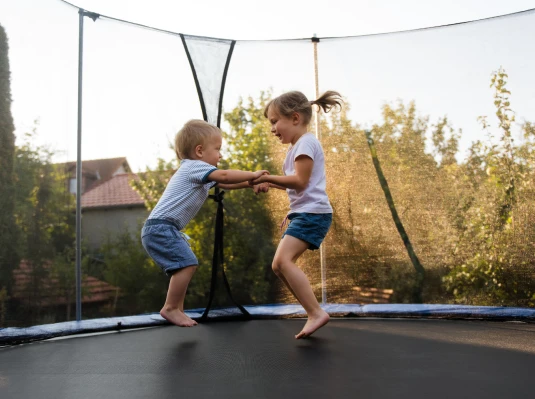 Two young children joyfully bounce on a trampoline, holding hands. They are surrounded by trees and houses on a sunny day, conveying a playful, carefree mood.