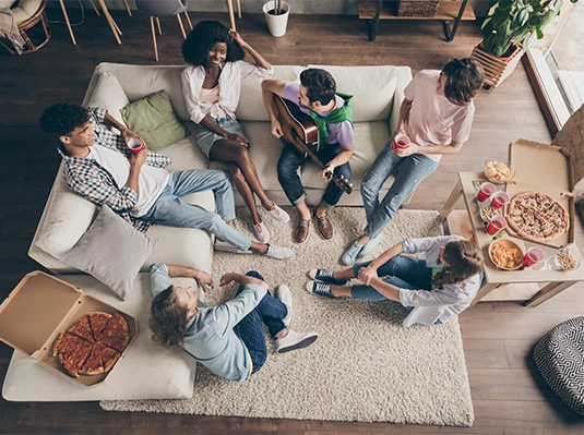 A group of friends relax in a cozy living room. They sit on a sofa and floor, eating pizza, drinking from red cups, and enjoying a guitar serenade.