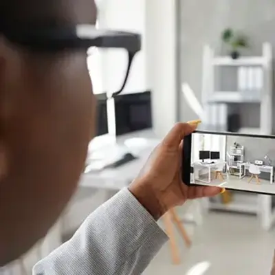 A person photographs a modern, minimalistic home office with a smartphone. The workspace features a desk, chair, plants, and shelves, conveying a tidy, organized feel.