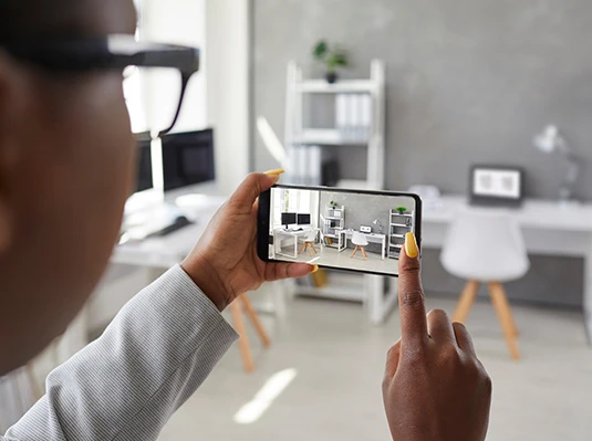 A person photographs a modern, minimalistic home office with a smartphone. The workspace features a desk, chair, plants, and shelves, conveying a tidy, organized feel.