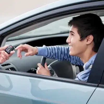 A smiling young man in a striped hoodie sits in a car, reaching for car keys handed to him through the window, conveying excitement and readiness.