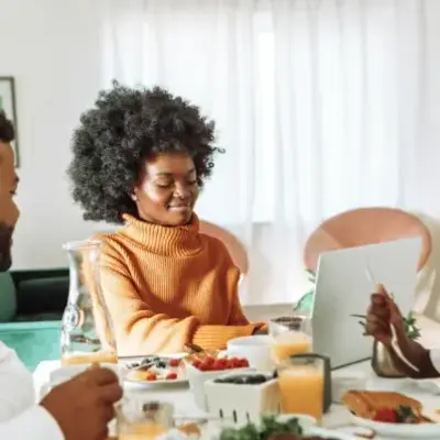 A family sits at a dining table with breakfast foods. A woman in an orange sweater uses a laptop, while a man and a child enjoy their meal. Bright, cozy atmosphere.