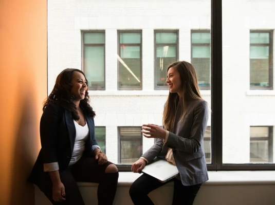 Two women engaged in conversation, standing by a window with natural light illuminating their discussion.
