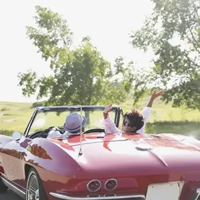A joyous couple enjoys a sunny drive in a vintage red convertible. The woman playfully raises her arm, surrounded by lush green trees and open fields.