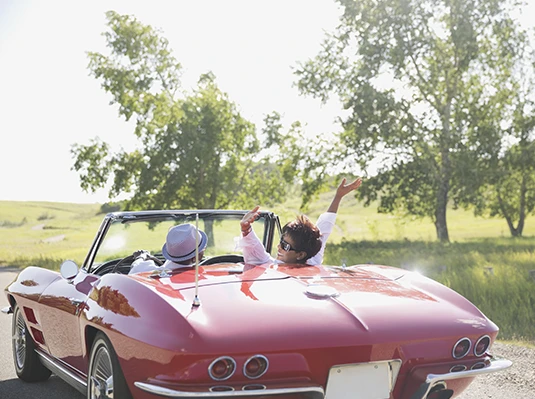A joyous couple enjoys a sunny drive in a vintage red convertible. The woman playfully raises her arm, surrounded by lush green trees and open fields.