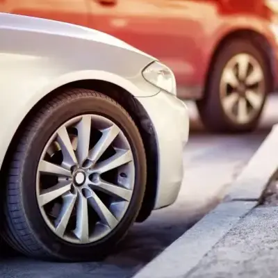 Close-up of a white car's front tire parked parallel to a curb. Two other cars are blurred in the background, creating a calm urban scene.