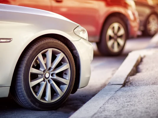 Close-up of a white car's front tire parked parallel to a curb. Two other cars are blurred in the background, creating a calm urban scene.